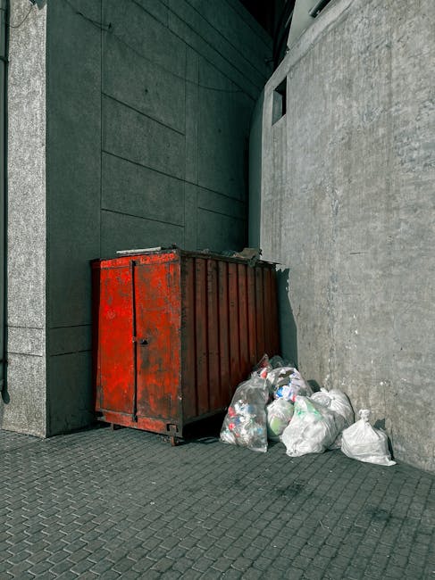 A red metal skip with a weathered, slightly rusted surface is placed against the corner junction of two grey building walls, one smooth and the other textured concrete. Adjacent to the skip, several c