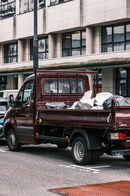 A small, dark red flatbed truck parked on a city street in front of a modern building with large, rectangular windows and a concrete facade. The truck's cargo bed is filled with various types of rubbi