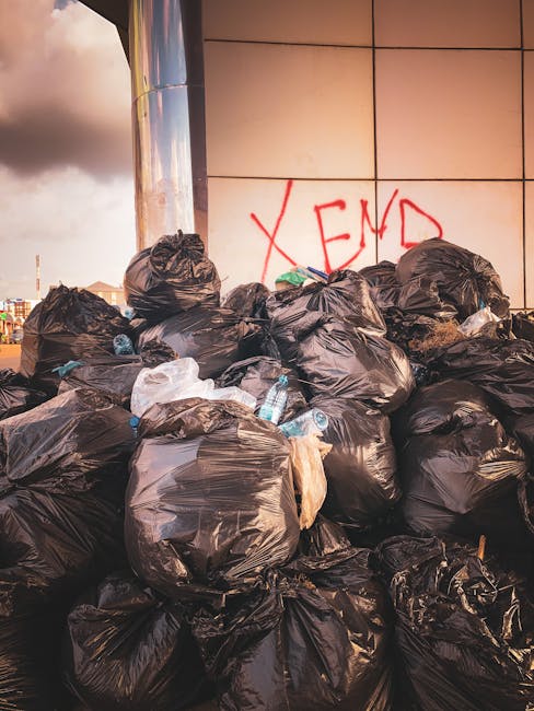 A large pile of black rubbish bags, some partially torn or bulging, is stacked against a modern building with a tiled exterior in an urban setting. Several clear plastic bottles are visible protruding