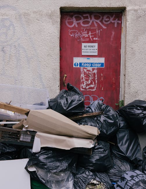 A red metal door with multiple graffiti tags in white and pink spray paint, situated within a beige, textured wall that appears weathered. The door features a white and blue 'Keep clear' sign and a sm
