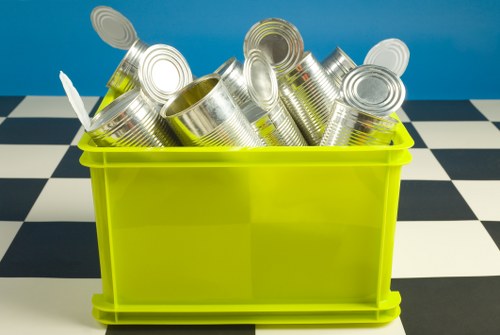 A bright yellow plastic recycling bin filled with numerous empty, silver tin cans featuring ridged sides and closed tops, some cans leaning and stacked atop each other inside the bin. The bin is positioned on a black and white checkered floor, with a solid blue background behind it. The scene is well-lit, emphasizing the metallic sheen of the cans and the vibrant colour of the bin, suggesting an indoor setting, possibly as part of a waste management or rubbish clearance operation in London. The image captures the contents clearly, highlighting the focus on metal recycling and the importance of rubbish removal services in urban environments like London, where Rubbish Clearances London operates.